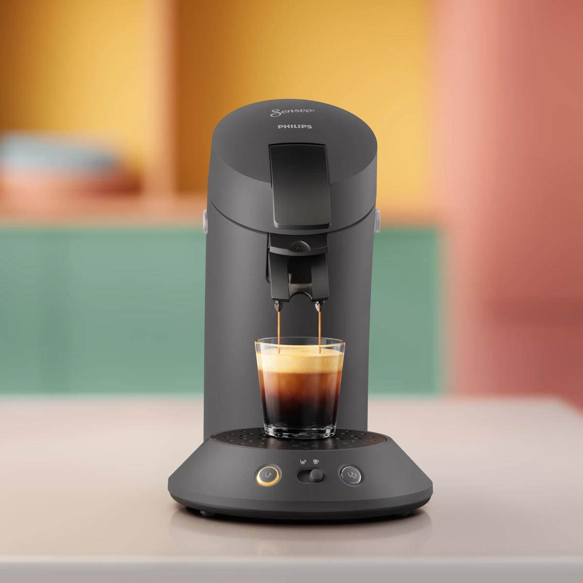 Una cafetera gris Philips Senseo prepara café en un vaso transparente con fondo colorido.