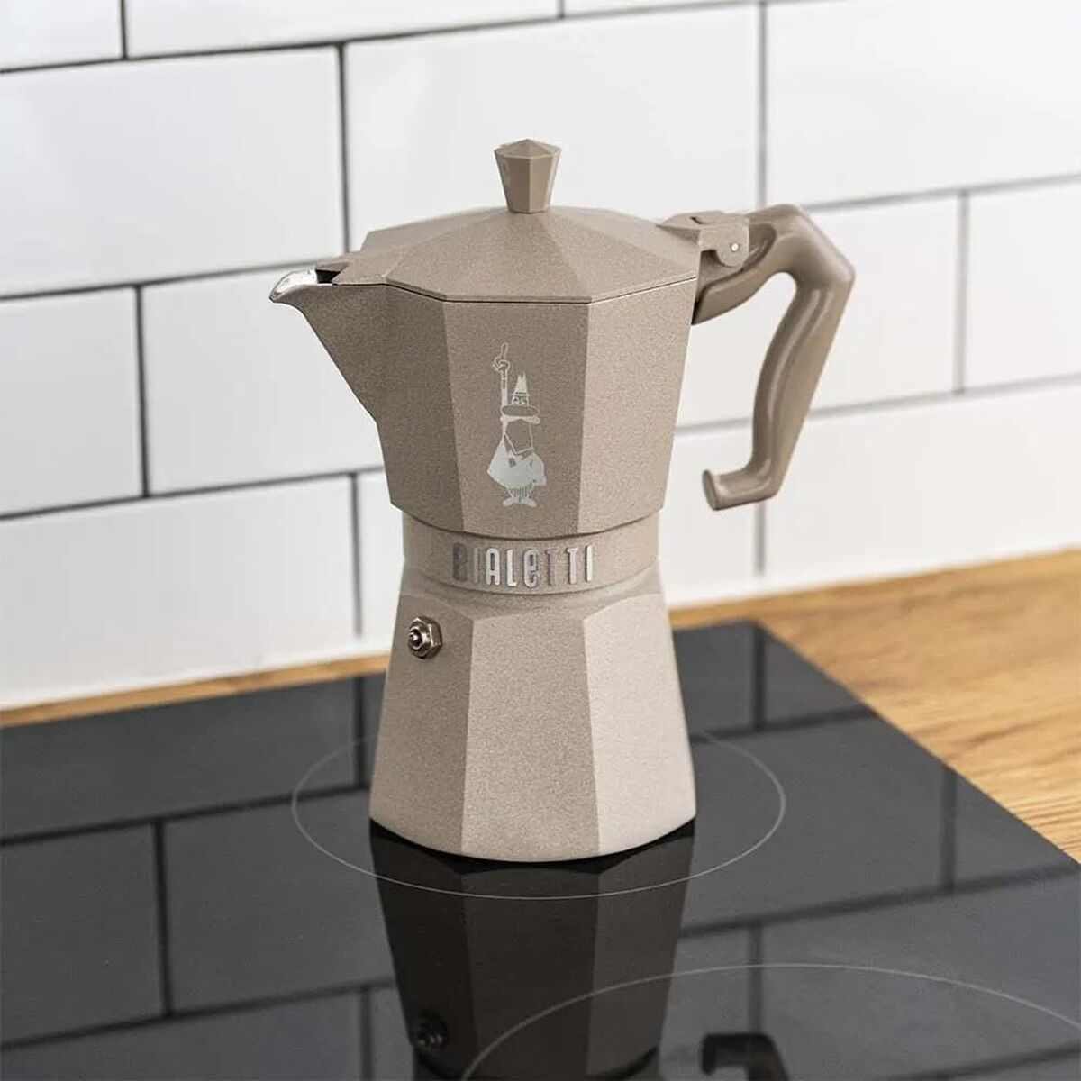 Una moka Bialetti color sabbia su un piano cottura a induzione nero e piastrelle bianche.