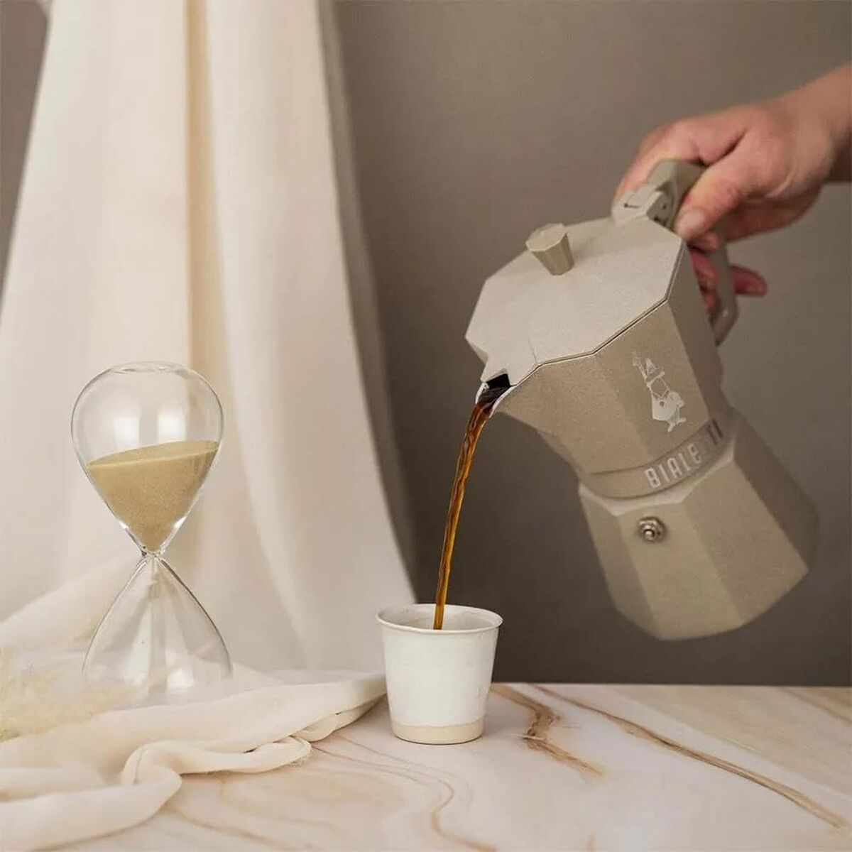 Una mano versa il caffè da una moka beige in una tazza. Una clessidra si trova accanto alla tazza.
