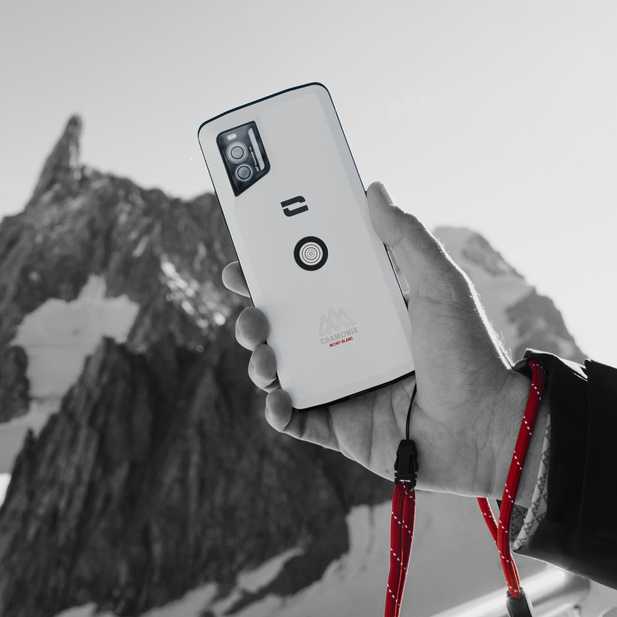 Ręka trzyma biały smartfon Chamonix na tle zaśnieżonych szczytów górskich na czarno-białym zdjęciu.