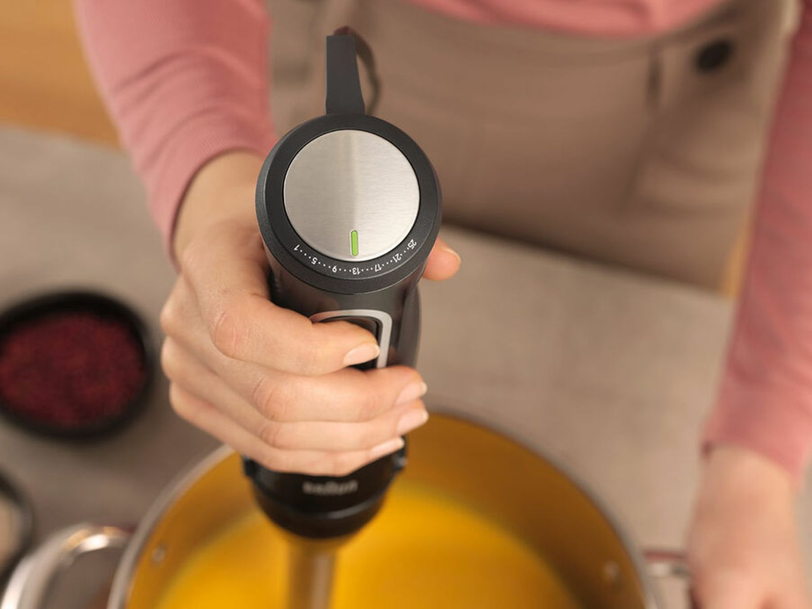 Persona sosteniendo una batidora de mano negra con selector de velocidad sobre una olla con sopa amarilla.