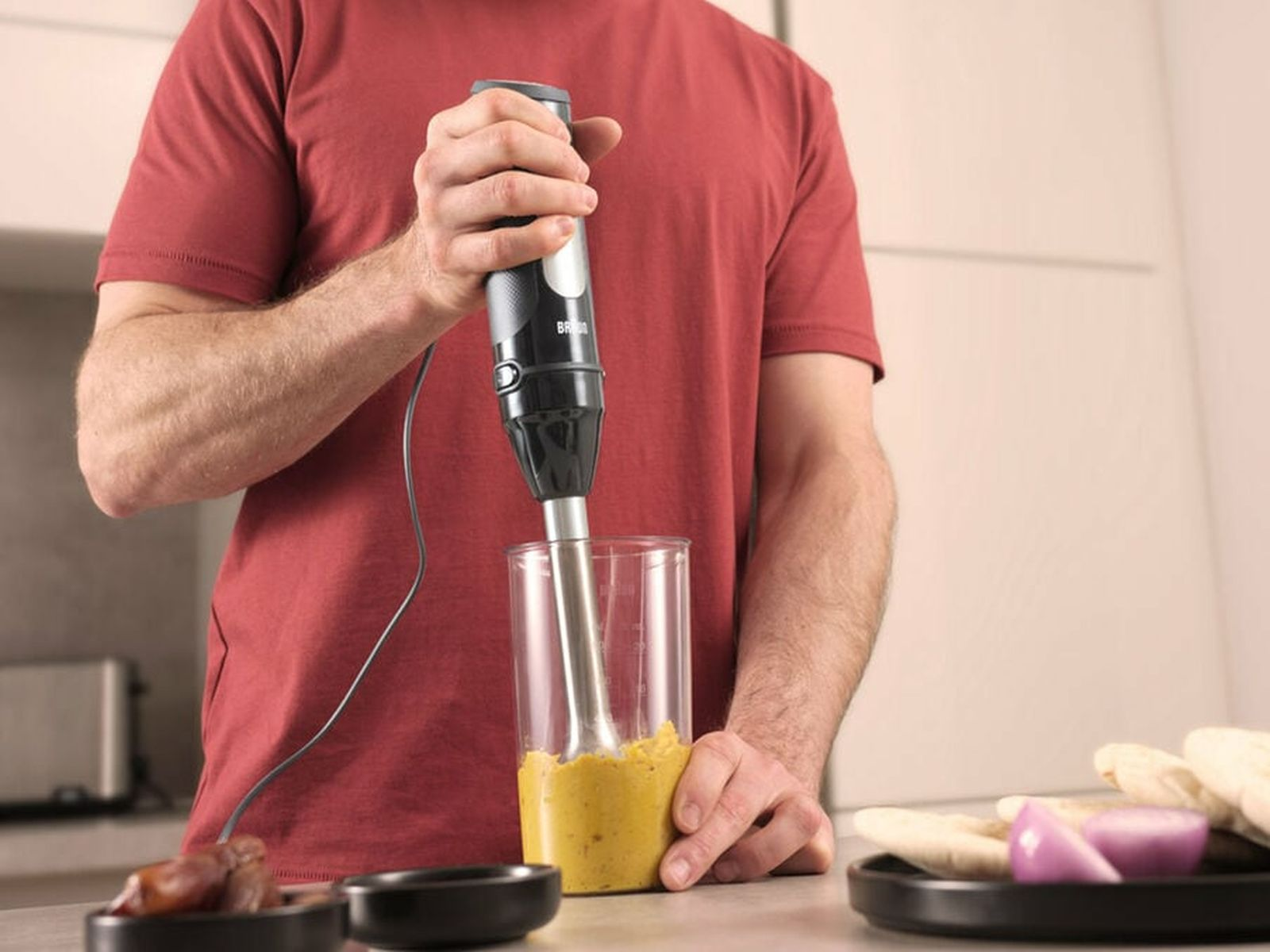 Un hombre con camisa roja sostiene una batidora de mano en un vaso con puré amarillo en una cocina.