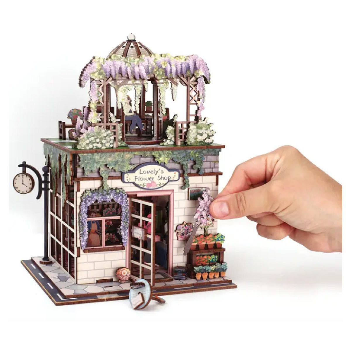 Hand setzt kleines Bauteil an detailreiches Modell eines Blumenladens mit Dachpavillon und Glyzinien.