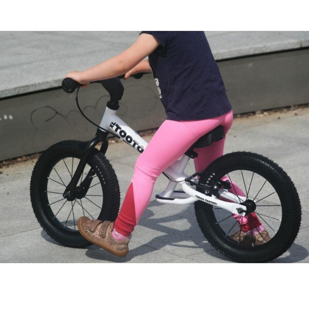 Ein kleines Kind in dunklem Hemd und rosa Leggings fährt auf gepflasterter Fläche ein Laufrad.