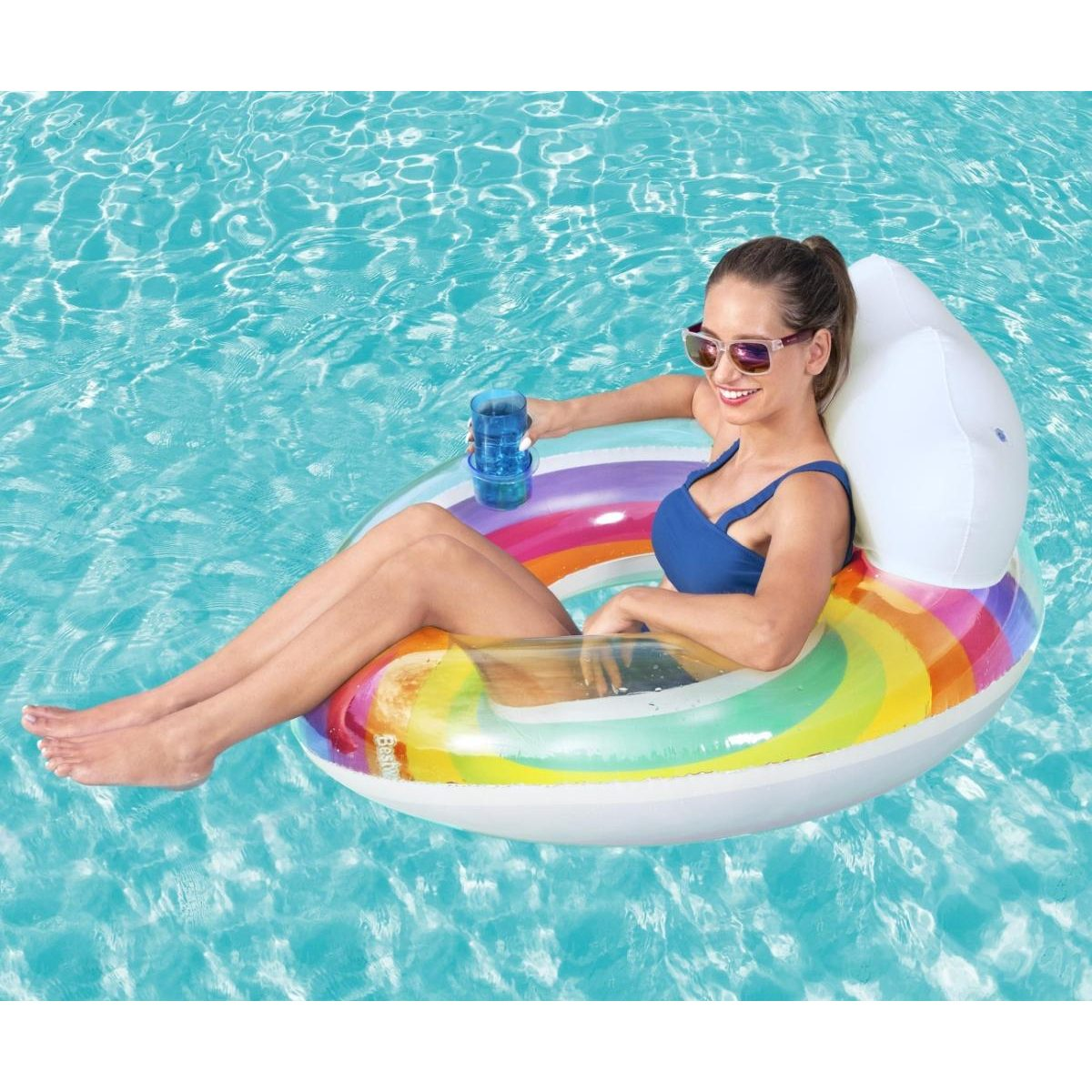 Una mujer se relaja en una piscina en un flotador inflable con rayas de arco iris y respaldo, sosteniendo un vaso azul.