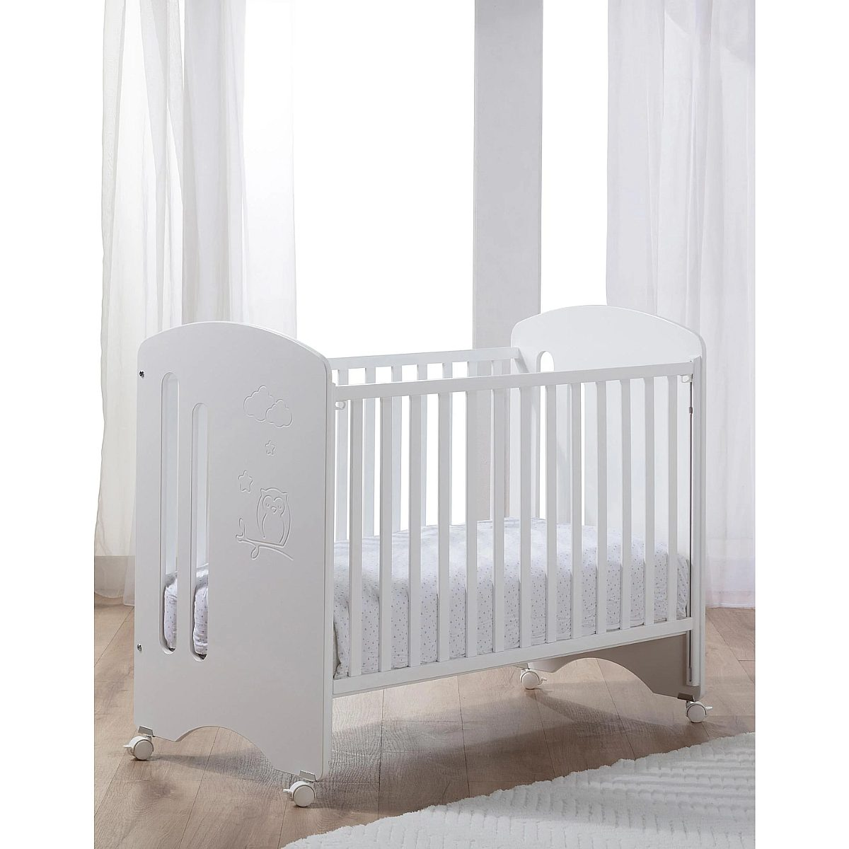 Ein weißes Babybett mit Rollen steht auf Holzboden vor einem Fenster mit weißen Vorhängen.