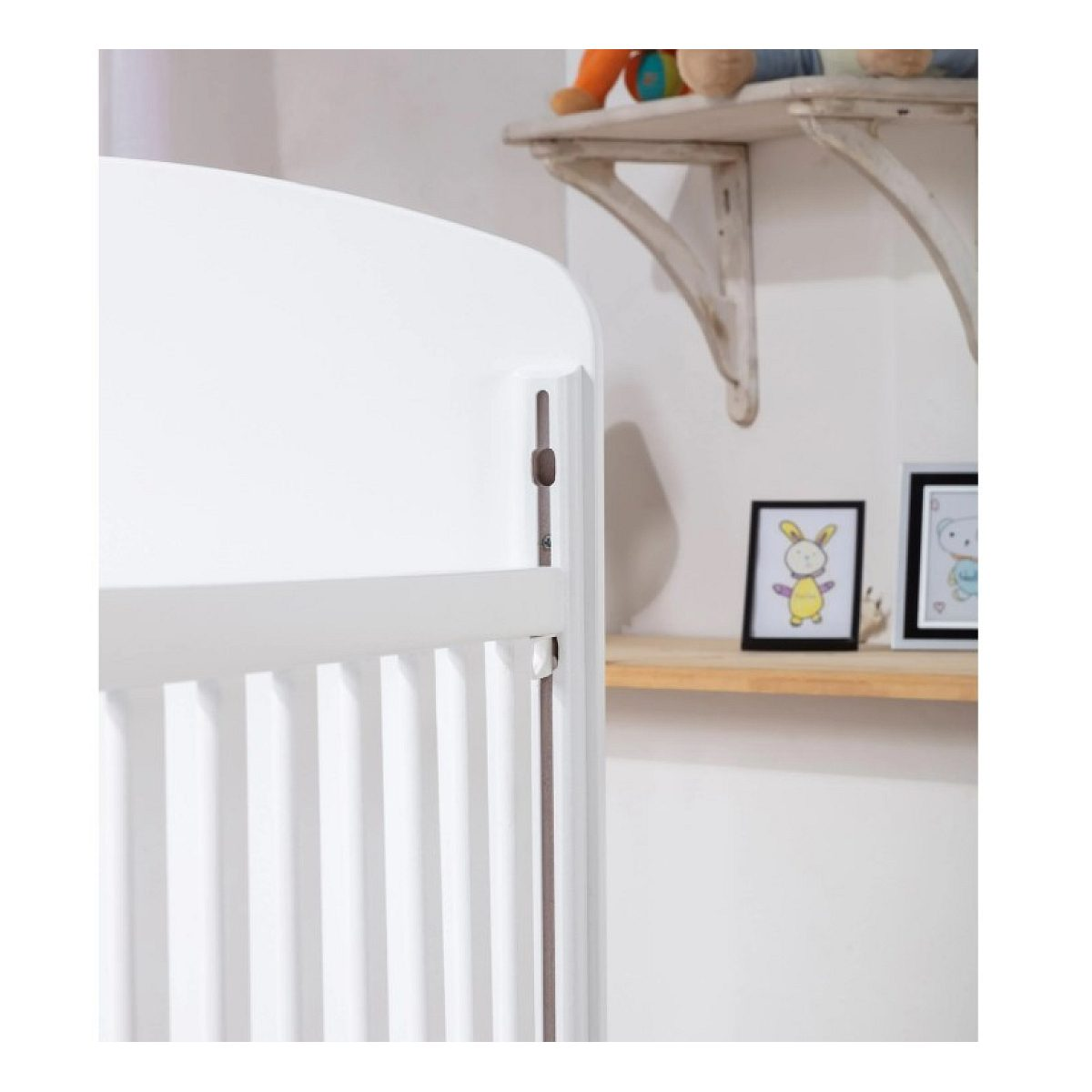 Weißes Babybett im Vordergrund, im Hintergrund ein Holzregal mit Bilderrahmen.