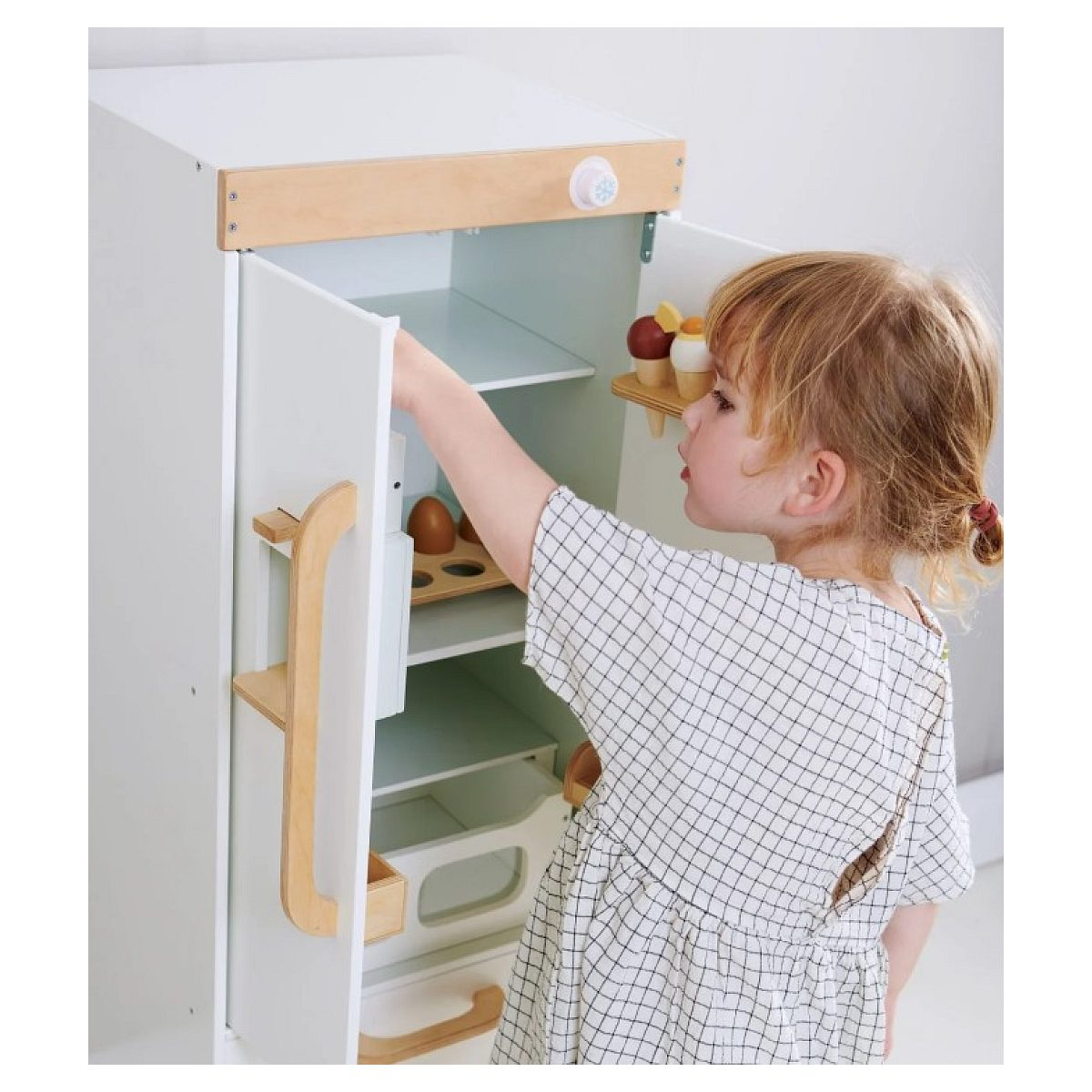 Ein kleines Mädchen im Karottenkleid öffnet einen weiß-hölzernen Spielzeugkühlschrank.