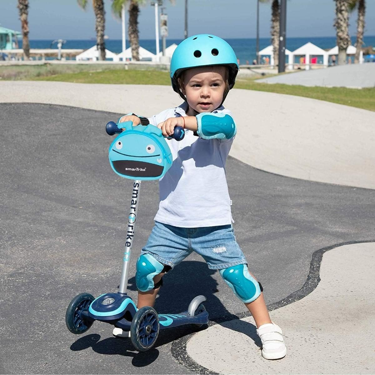 Kleinkind mit blauem Helm und Schutzkleidung fährt Roller im Park am Meer.