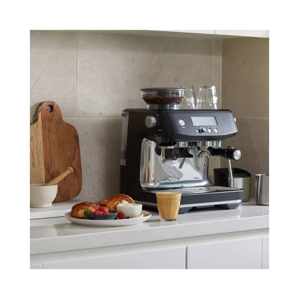 Une machine à espresso noire sur un comptoir de cuisine avec des pâtisseries, des fruits et une planche à découper.