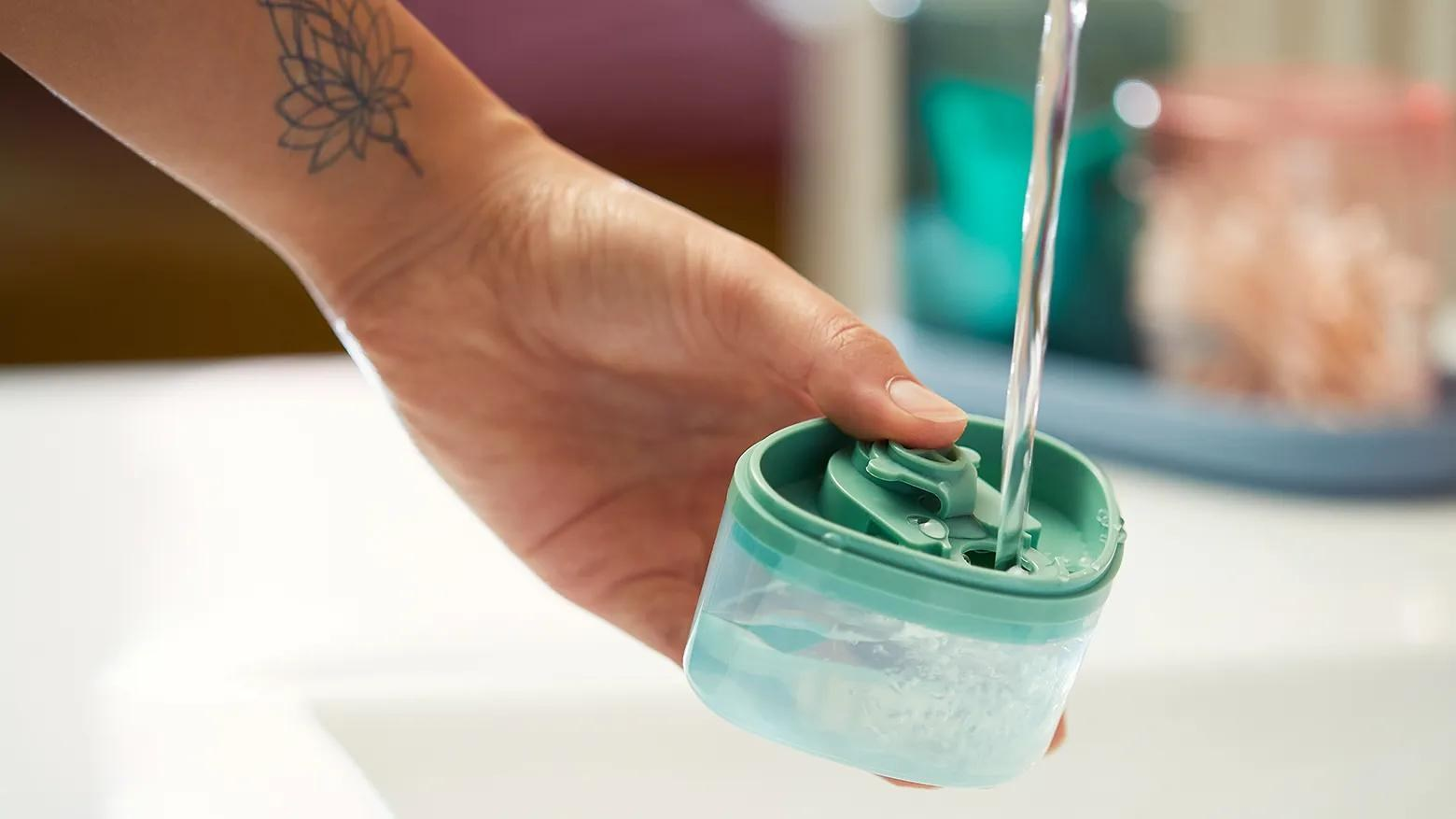 Eine tattoowierte Hand hält einen kleinen, grünen Behälter, während Wasser hineinfließt.
