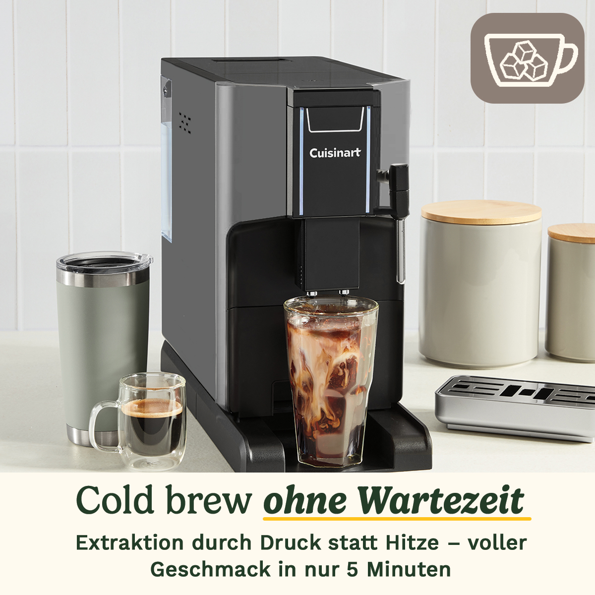 Una cafetera Cuisinart gris prepara café frío en un vaso, flanqueada por tazas y botes.