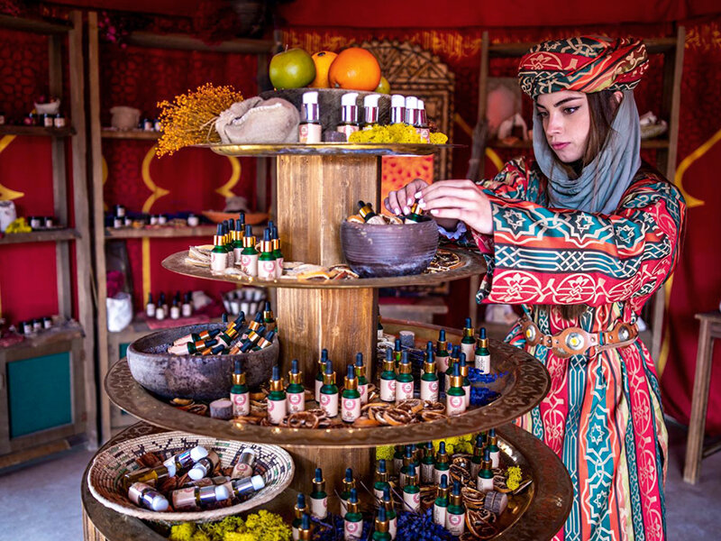 Una mujer con traje tradicional organiza botellas pequeñas en un soporte de madera con aceites y frutas.