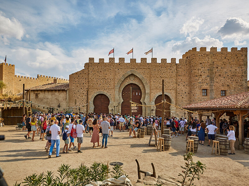 Una gran multitud de visitantes se reúne frente a una fortaleza de piedra medieval con puertas de madera y banderas.