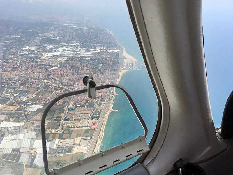Vista desde la ventana de un avión sobre una ciudad costera con playa y mar azul.