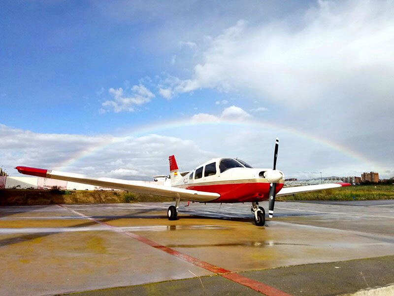 Un avión de hélice rojo y blanco aparcado en asfalto mojado bajo un cielo con un arco iris.