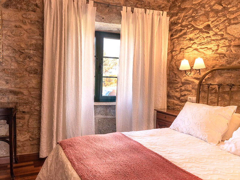 Un dormitorio rústico con paredes de piedra, cama con ropa blanca y roja, cortinas blancas y lámparas de pared.