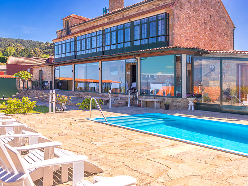 Un hotel de piedra con grandes ventanales de vidrio cuenta con una piscina azul brillante en un patio con tumbonas blancas.