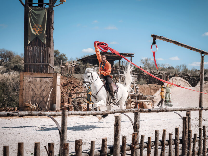 Un jinete en un caballo blanco sostiene una bandera roja en una arena con torres de madera.