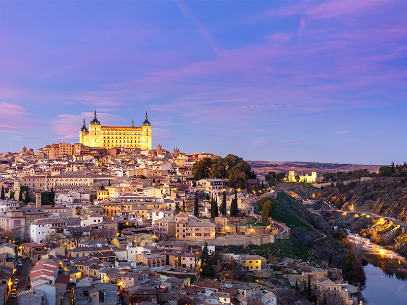 Ciudad histórica iluminada con castillo en la colina, río y cielo crepuscular púrpura.