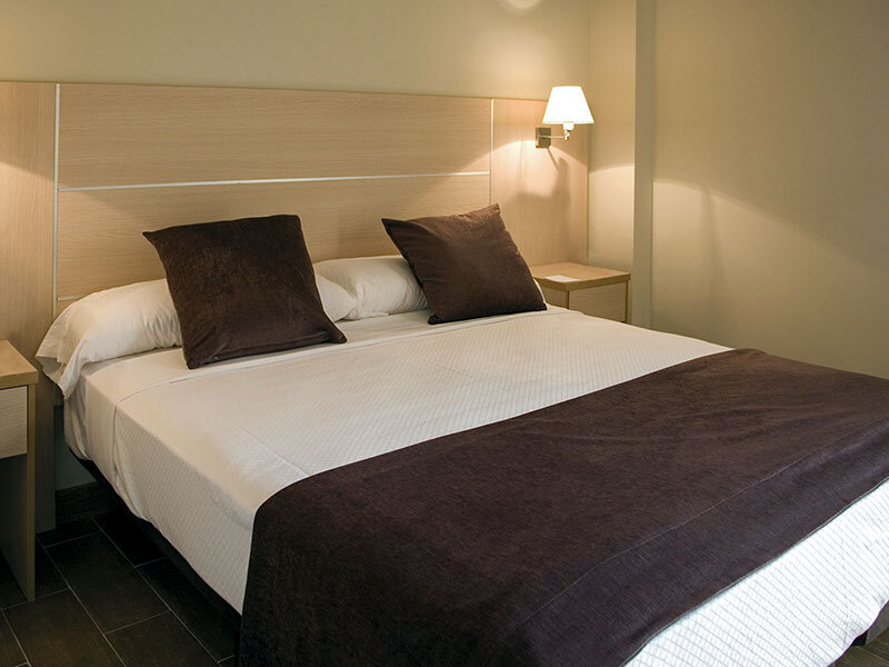 Una habitación de hotel moderna con una cama grande, ropa de cama blanca, cojines marrones y un cabecero de madera.