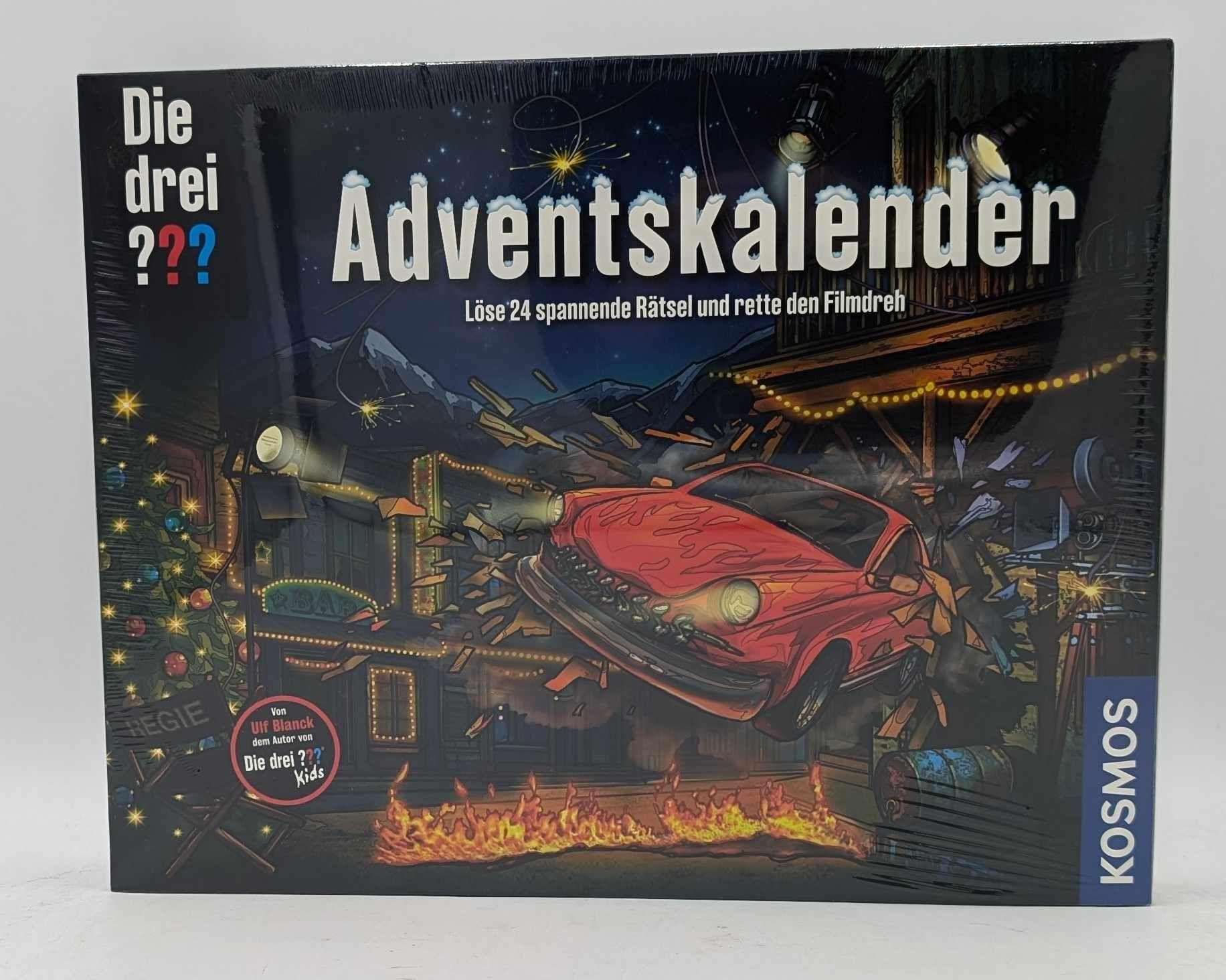 Ein Brettspiel-Cover zeigt ein rotes Auto, das durch ein Filmset kracht, mit Flammen am Boden und dem Titel Adventskalender.