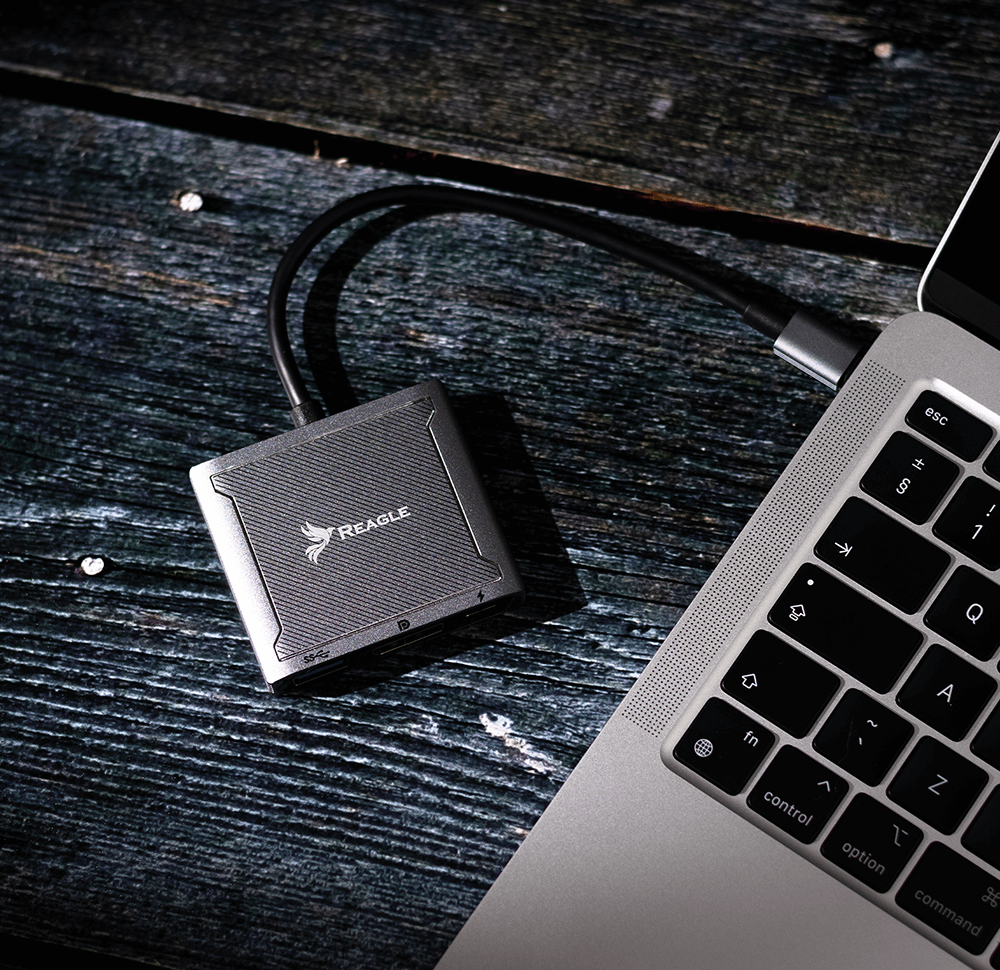 Ein grauer Reagle USB-Hub ist über ein Kabel mit einer silbernen Laptoptastatur auf dunklem Holz verbunden.