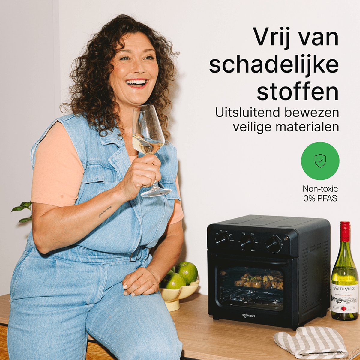 Smiling woman in denim overalls holds a wine glass next to a black air fryer and a wine bottle on a counter.