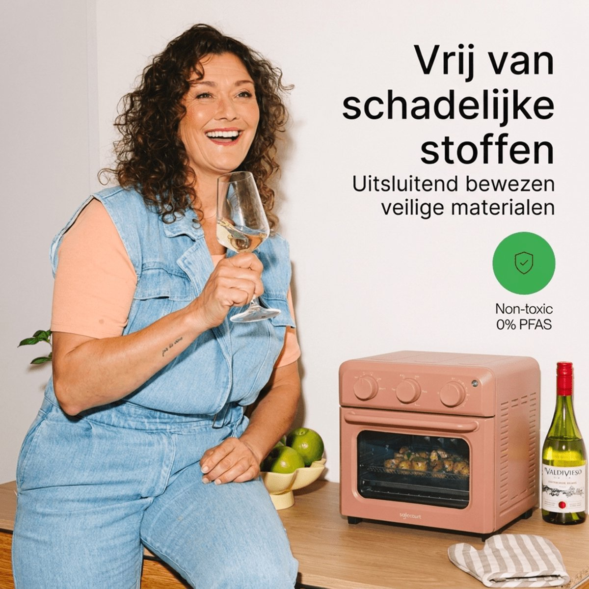 Smiling woman holds wine glass near a pink air fryer and wine bottle on a counter, with Dutch text overlay.