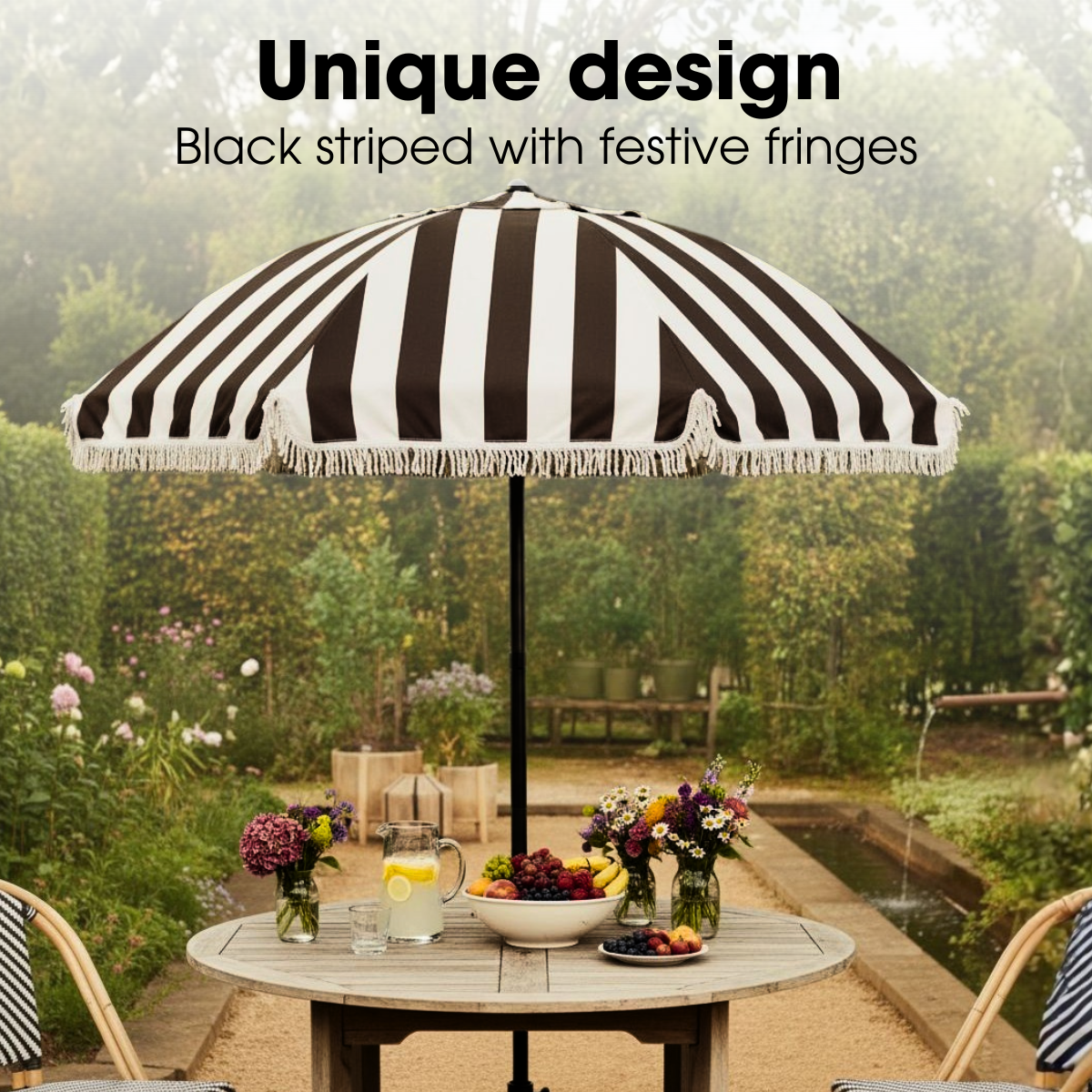 A black and white striped patio umbrella with fringes stands over a wooden table set with food and flowers in a garden.