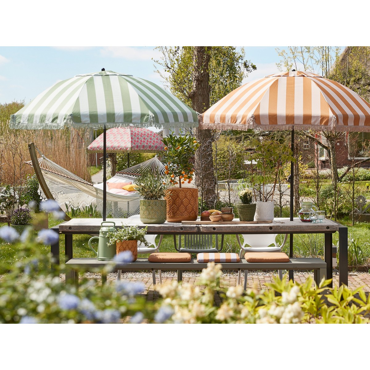 Garden table with two large striped umbrellas in green and orange, plants, and a hammock in the background.