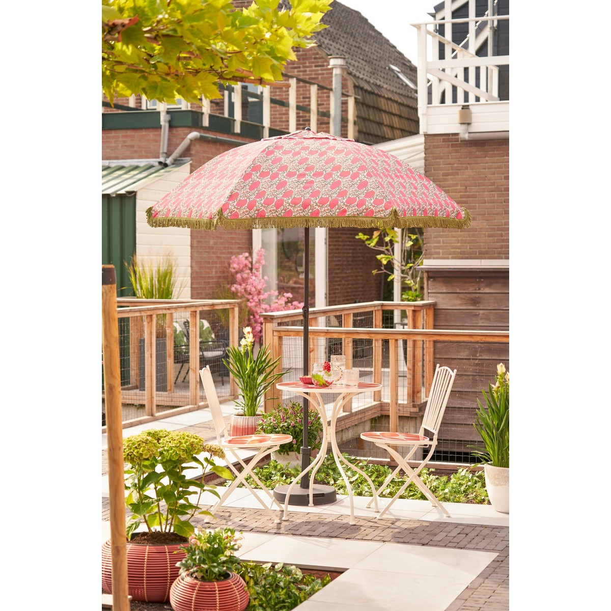 A patio features a round table and two chairs under a pink patterned umbrella, surrounded by potted plants.