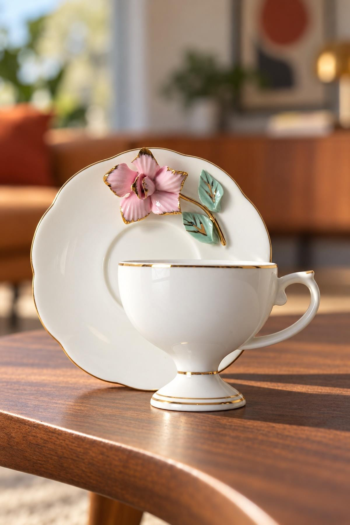 Altın kenarlı beyaz porselen çay fincanı, pembe bir çiçekle süslenmiş uyumlu bir tabağın önünde duruyor.