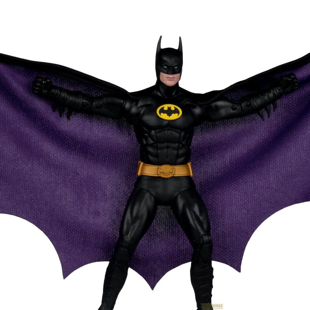 Un action figure di Batman nera con cintura gialla e un grande mantello viola esteso su sfondo bianco.