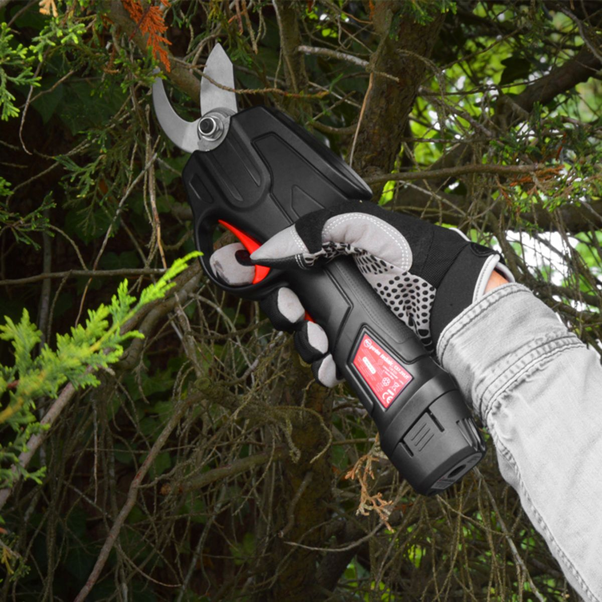 Eine Hand im Arbeitshandschuh hält eine elektrische Gartenschere vor Heckensträuchern.