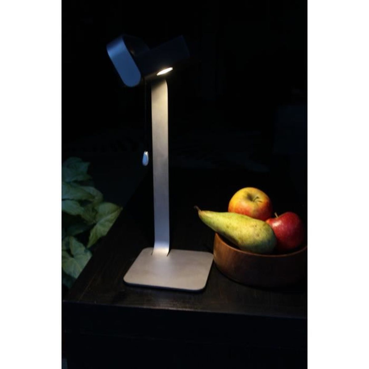 Eine silberne Schreibtischlampe beleuchtet eine Holzschale mit Äpfeln und einer Birne auf dunklem Tisch.