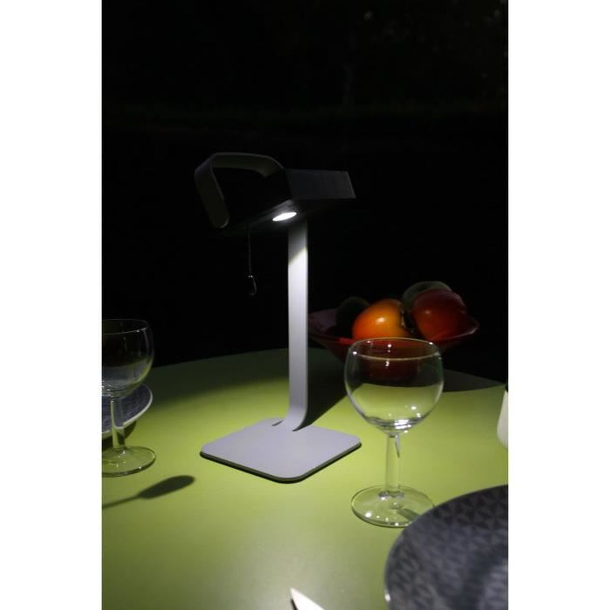 Eine weiße Schreibtischlampe beleuchtet einen grünen Tisch mit Weingläsern und einer Obstschale nachts.