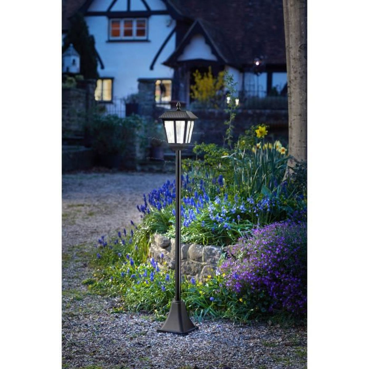 Eine schwarze Gartenleuchte steht abends in einem Garten vor einem beleuchteten Haus.