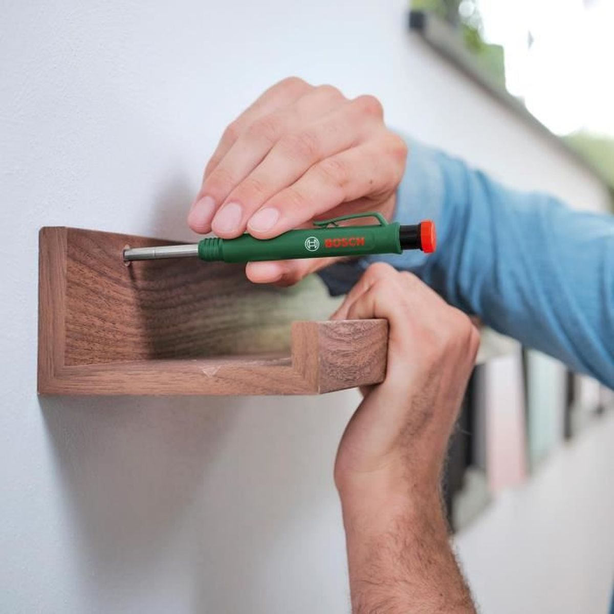 Eine Person benutzt ein grünes Bosch-Werkzeug, um ein Holzregal an einer weißen Wand zu montieren.