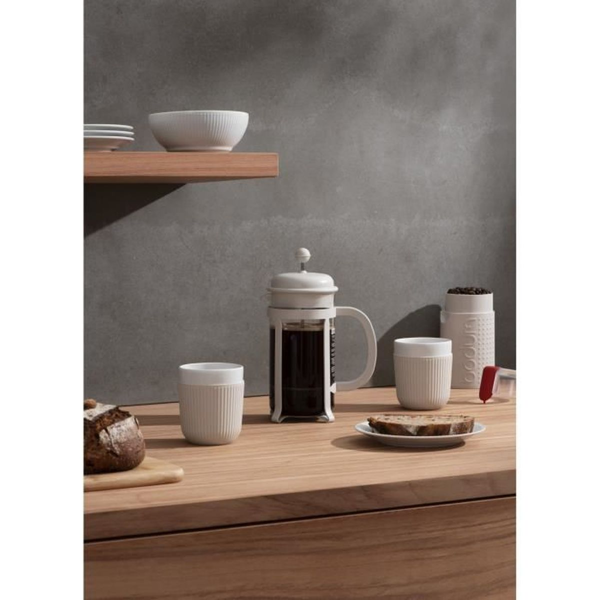 Weiße Kaffeepresse mit zwei Tassen und Brot auf Holztheke vor grauem Hintergrund.