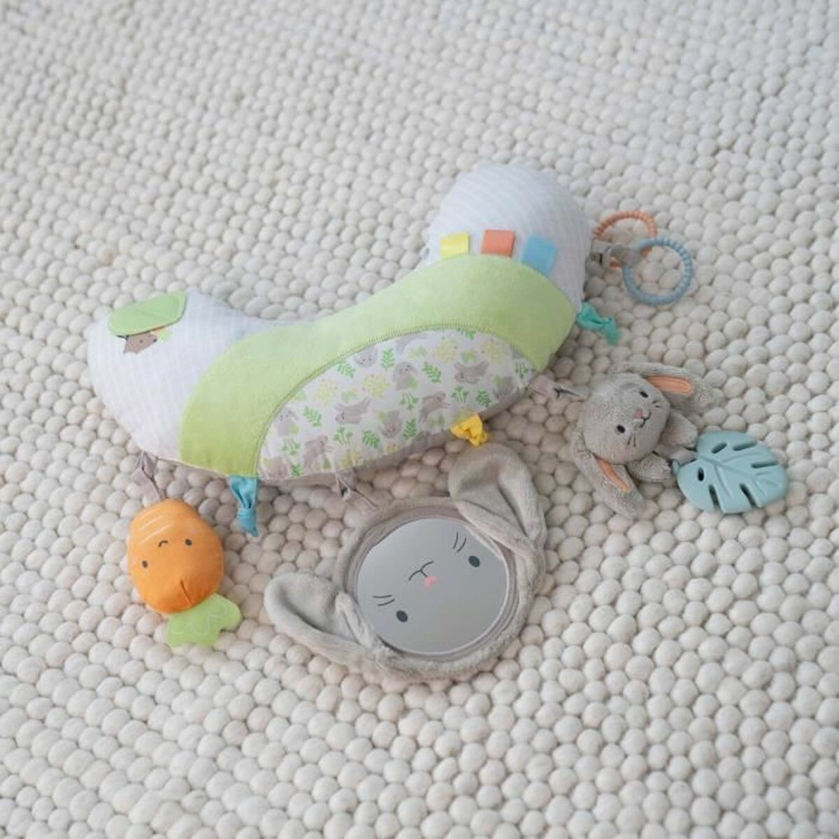Ein weiß-grünes Babyspielkissen mit Hase, Karotte, Spiegel und Rassel auf hellem Teppich.
