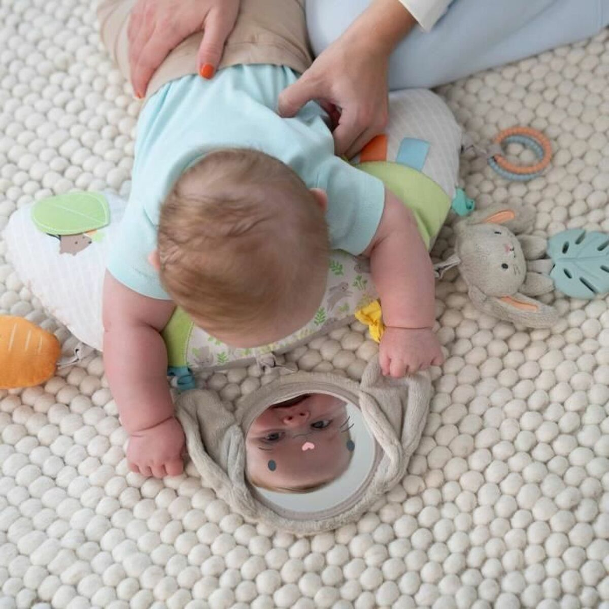 Ein Baby liegt auf einem gemusterten Kissen auf einem weißen Teppich und blickt in einen Plüschspiegel.