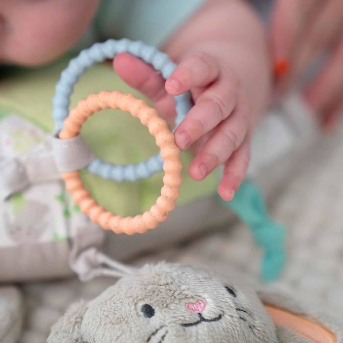 Eine Babyhand greift nach orangefarbenen und blauen Beissringen ueber einem Plueschtiere.