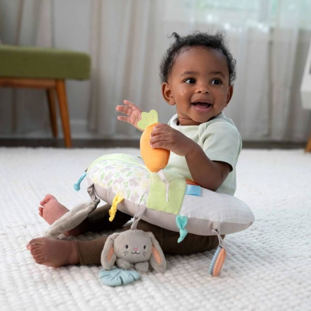 Ein lächelndes Baby sitzt auf einer weißen Matte, hält ein Orangenspielzeug und wird von Kissen umgeben.