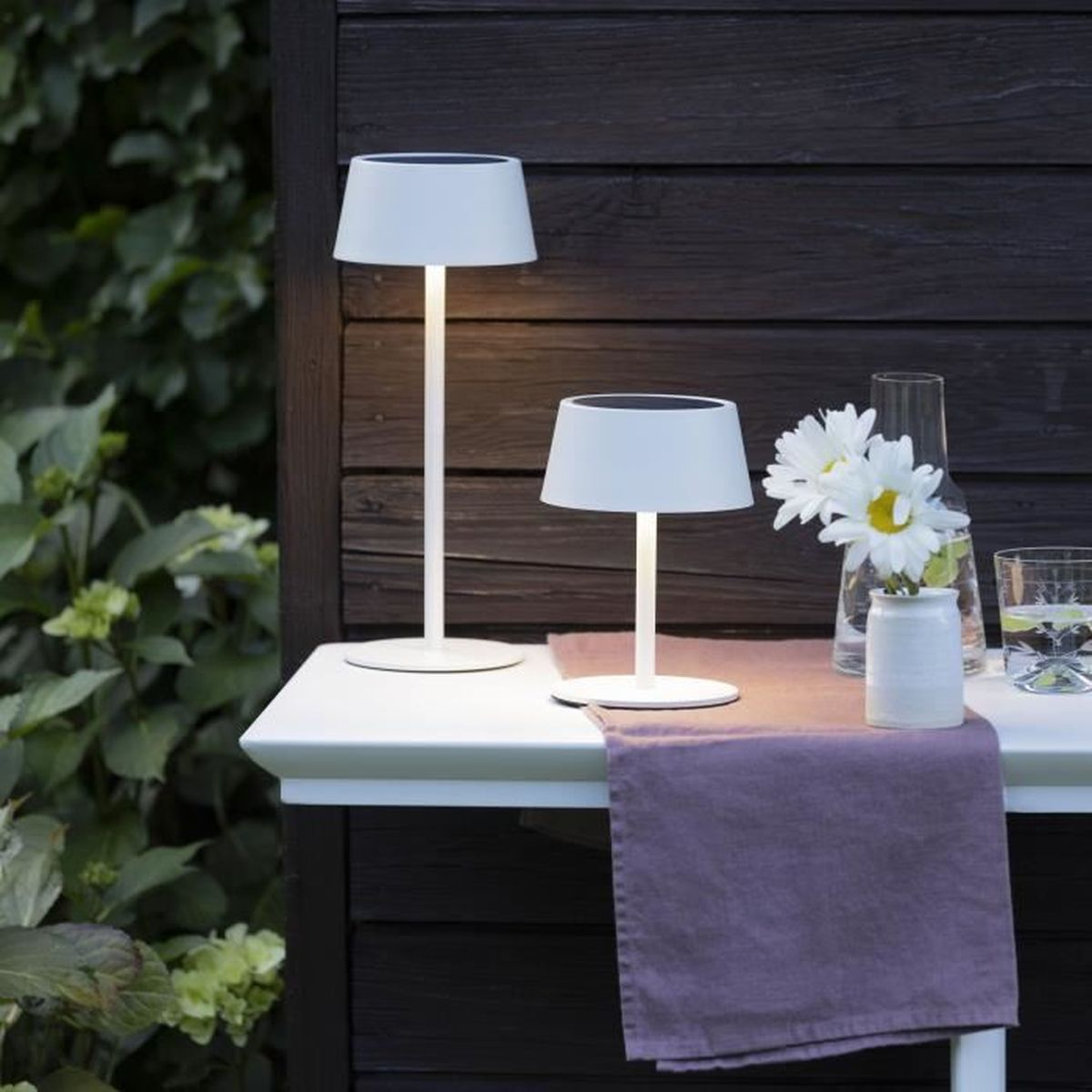 Zwei weiße Solarleuchten und eine kleine Blumenvase stehen auf einem weißen Gartentisch mit lila Läufer.