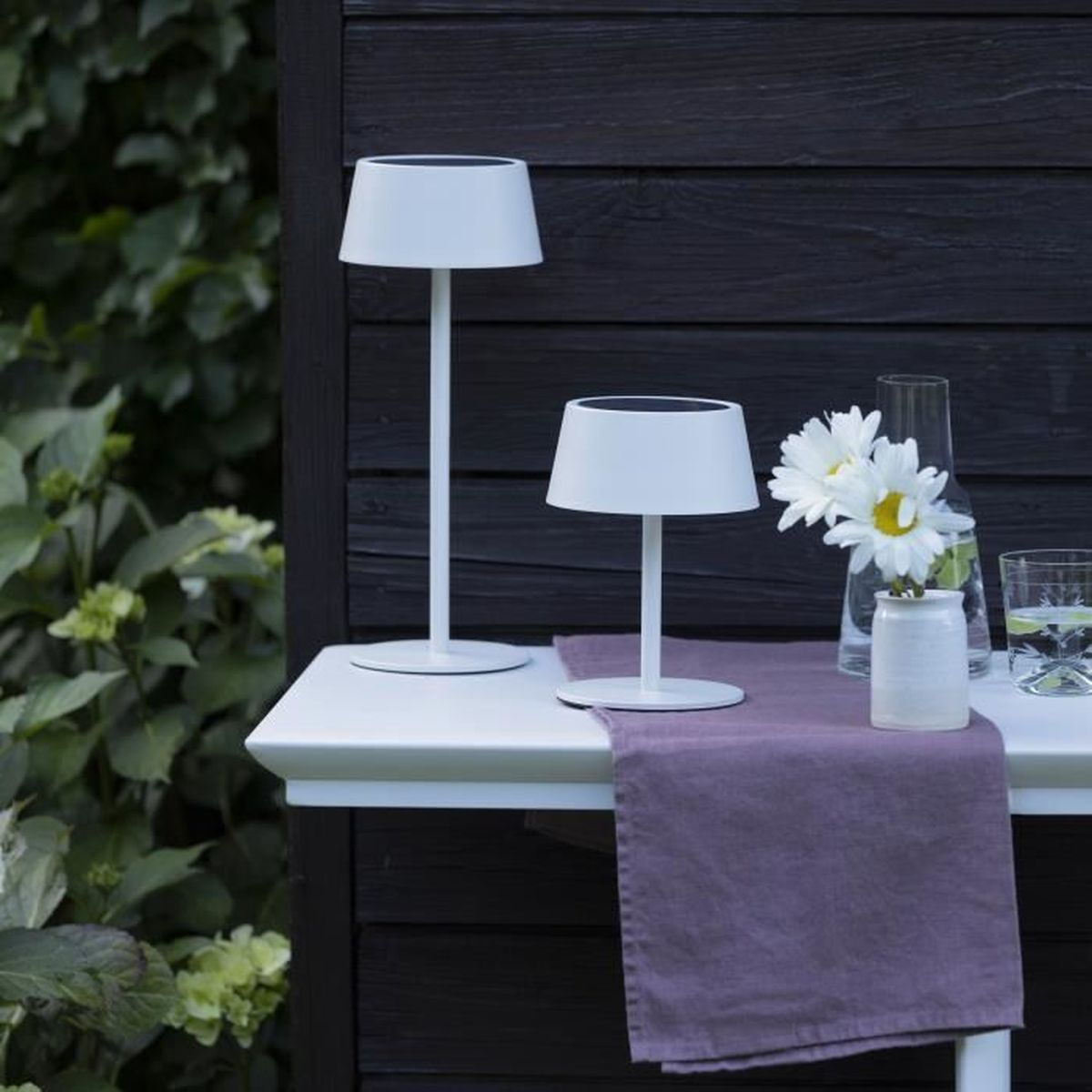 Zwei weiße Solarleuchten, weiße Blumen in einer Vase und Gläser stehen auf einem Tisch vor schwarzem Holz.