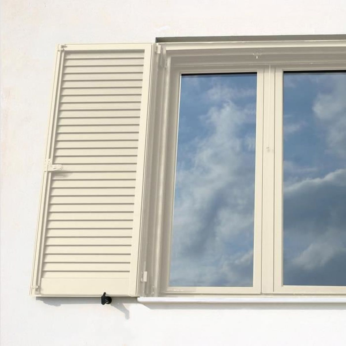 Ein beige Fenster mit einem offenen Lamellenladen an einer weißen Wand zeigt den blauen Himmel.