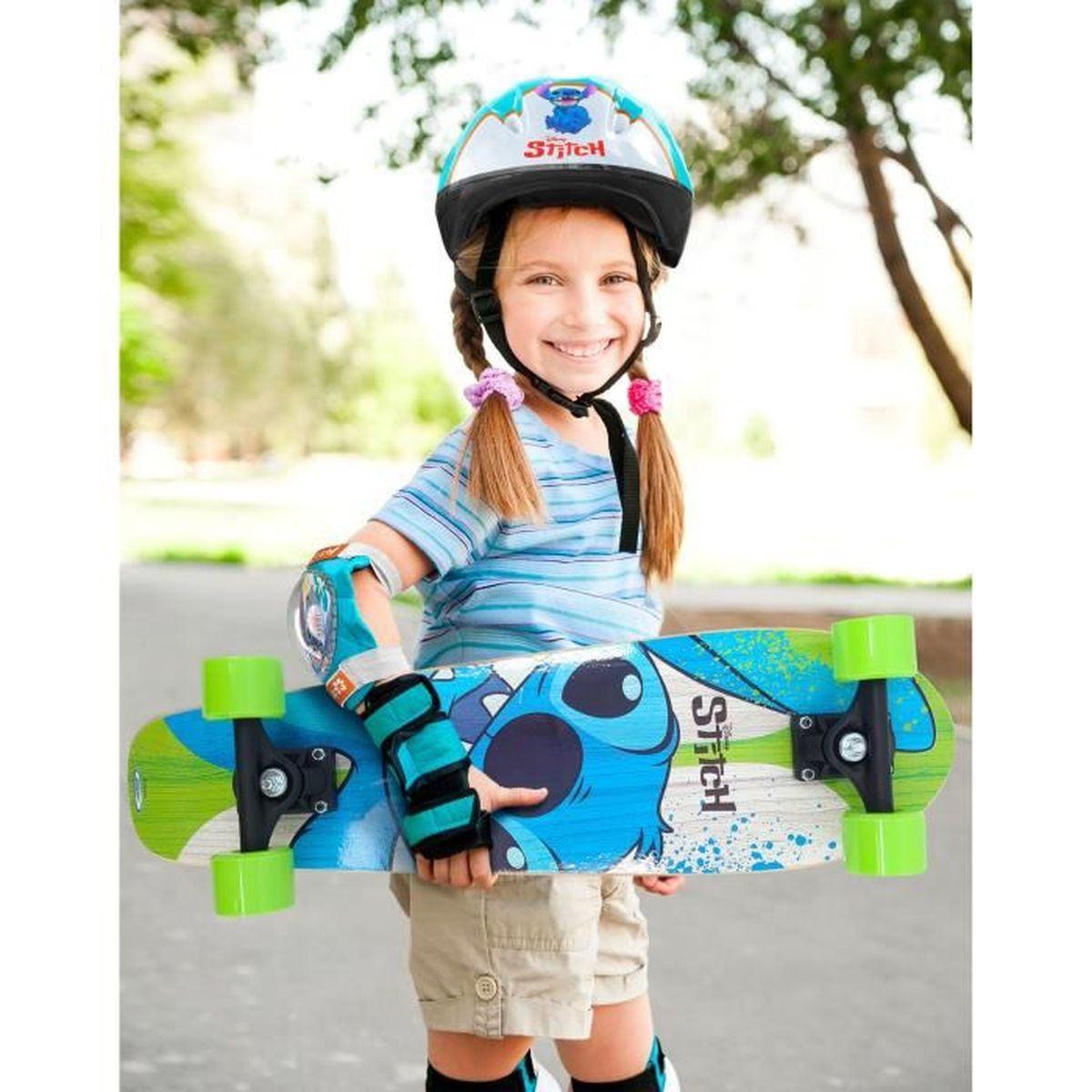 Ein junges Mädchen lächelt und hält ein Stitch-Skateboard, trägt Schutzausrüstung wie Helm, Ellenbogen- und Knieschoner.