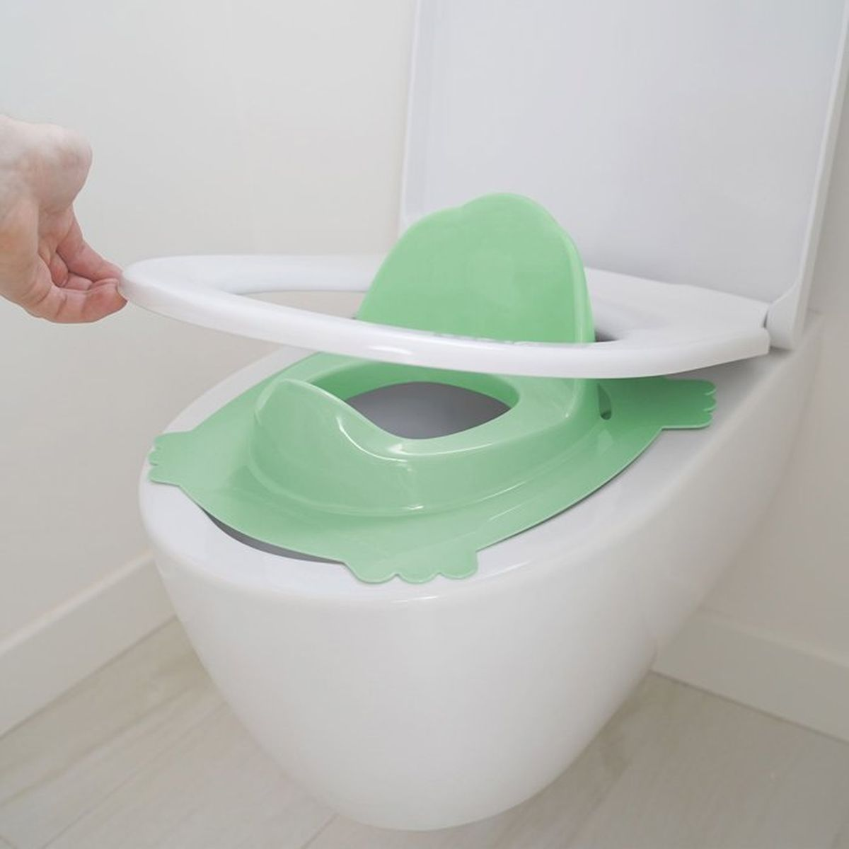 Eine Hand hält einen weißen Toilettensitz über einem grünen Kindersitz auf einer weißen Toilette.