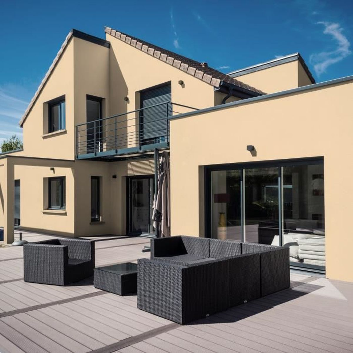 Moderne gelbe Villa mit Terrasse, dunklen Rattanmöbeln und blauem Himmel im Hintergrund.